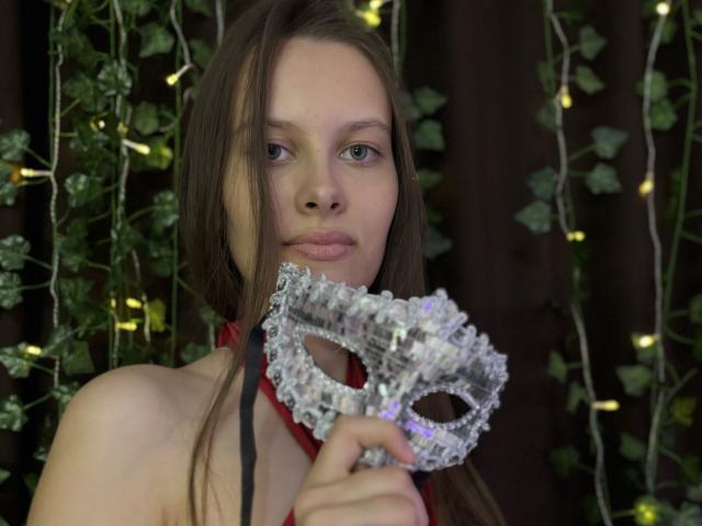 AyumiKirk with long brown hair holding a sparkly silver masquerade mask in front of her face, wearing a red halter, looking serious with green eyes.