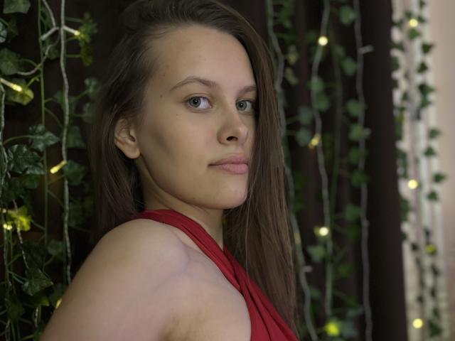 AyumiKirk with light skin, brown hair, green eyes, and red dress, looking over shoulder with a subtle smile, showing small tits and a slight armpit.