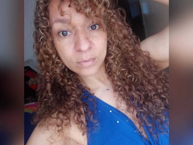 Close-up of Angelonix with curly, medium-brown hair and blue eyes. Angelonix wears a blue, sleeveless top with small, silver studs. Angelonix's right arm is raised, resting behind their head. Skin is fair with visible freckles. The expression is neutral, slightly pouting. The lighting is even, highlighting the texture of the hair and the fabric of the top. The image is focused on Angelonix's face and upper torso.