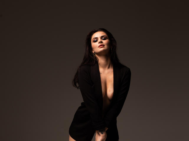 MilashaLovely with dark hair, wearing a black open shirt, showing her tits, standing against a dark background, looking up with a slight smile, hands near her crotch.