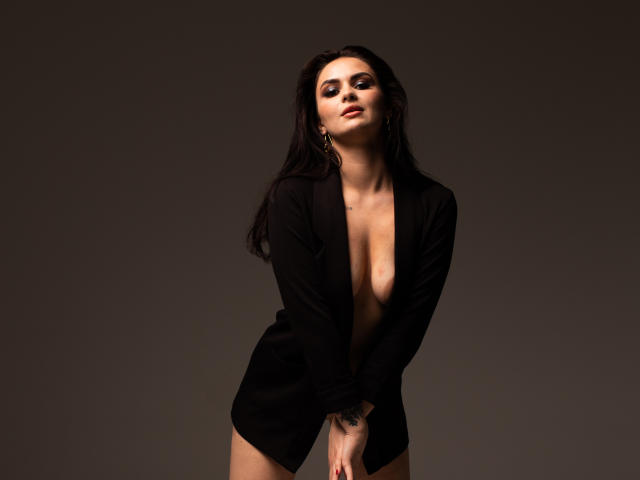 MilashaLovely with long black hair, wearing an open black shirt, exposing her tits, standing against a dark background, looking down with a slight smile, hands near her crotch.