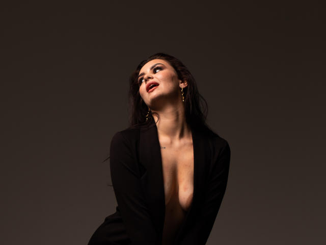 MilashaLovely with dark hair, wearing a black jacket, exposing her tits, looking up with red lips, gold earrings, against a dark background. Sheer, sexy, and confident.