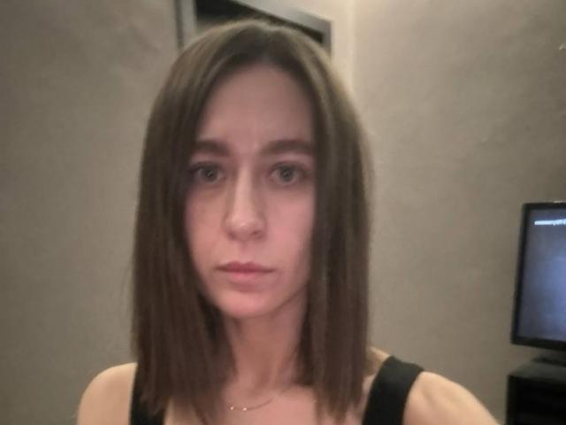 Sanezhka has straight brown hair, a pale face, and a neutral expression. She's wearing a black sleeveless top with visible straps. Her ass is not visible.