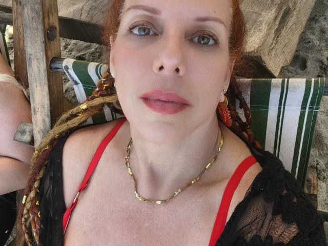Khharha's face, pale skin, red bra, gold chain necklace, black lace top, red lips, braided hair, green-striped chair, wooden frame, slight tan lines, relaxed expression, subtle makeup.