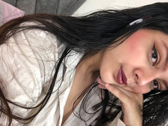 Violetta Rouse, con lunghi capelli neri, guance rosate, rossetto rosa scuro, camicia bianca, una mano sotto il mento, che guarda la fotocamera, luce soffusa, espressione rilassata..