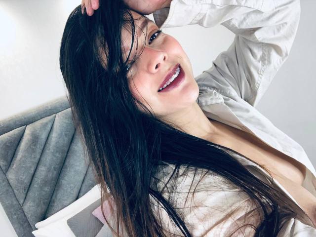 ViolettaRouse with long black hair, smiling, wearing an open white shirt, hand in hair, visible cleavage, sitting on gray upholstered headboard, soft white background, slightly tilted.