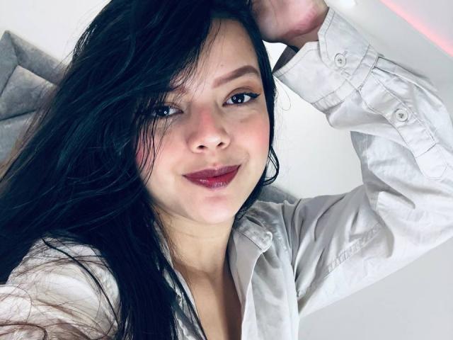 ViolettaRouse with long black hair, white button-up shirt, dark pink lipstick, and a slight smile, hand in hair, looking at camera, soft light, slightly flushed cheeks.