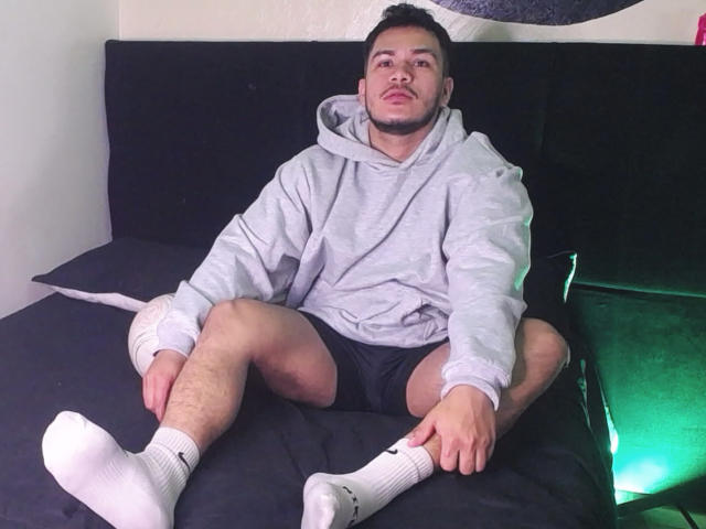 JeisonStark sits on a bed, legs spread, wearing a gray hoodie, black shorts, and white socks. His short hair and stubble are visible. His ass is slightly exposed.