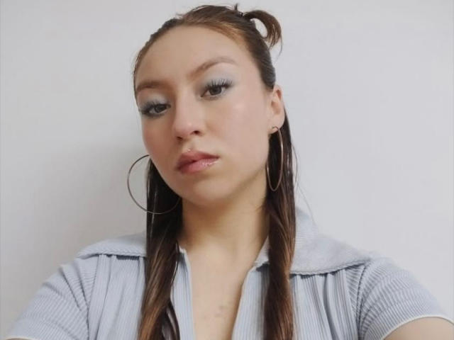 BellaLuck with shiny blue eyeshadow, small hoop earrings, and wet brown hair in a ponytail, wearing a gray ribbed shirt, looking slightly bored.