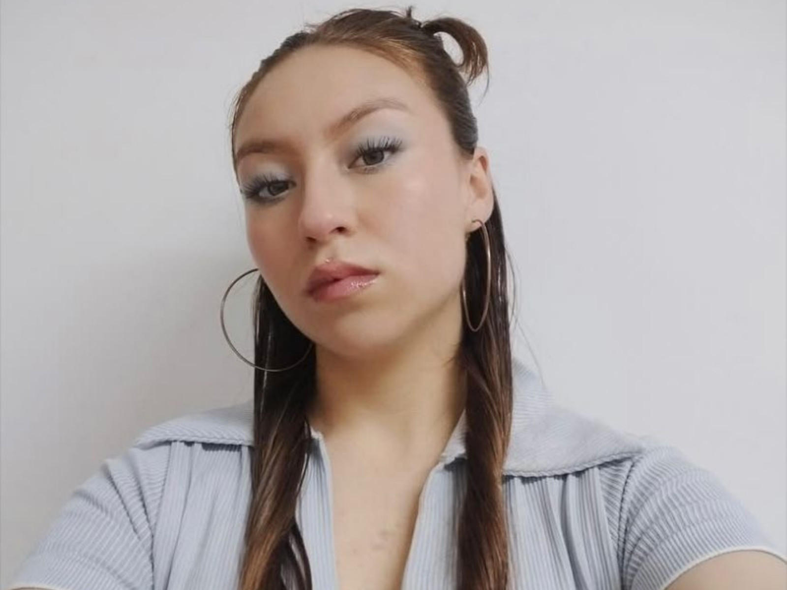 BellaLuck with shiny blue eyeshadow, small hoop earrings, and wet brown hair in a ponytail, wearing a gray ribbed shirt, looking slightly bored.