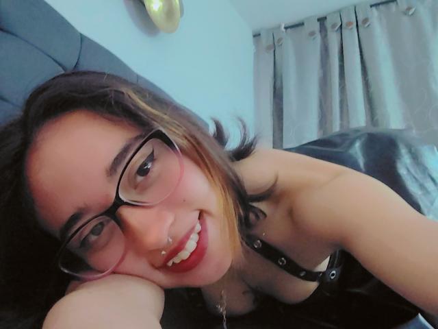 CamiVargas with glasses, nose ring, and red lipstick, smiling at the camera, lying on a bed, wearing a black strap top, showing her tits and ass.