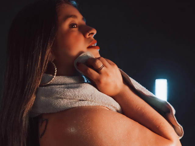 MelindaBoobs with wet skin, holding a towel over her tits, brown eyes, long dark hair, silver hoop earrings, and a tattoo on her arm, illuminated by a light source.