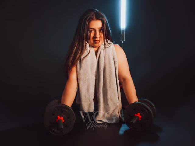 MelindaBoobs with long brown hair, wearing a gray towel, holding black dumbbells with red clips, in a dimly lit gym, looking determined. Bright light behind.