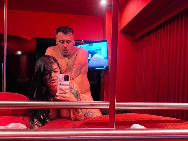 JackAndTriana, with black hair and large tits, on red bed, being fucked from behind by a tattooed guy with a serious face, taking a selfie in a red room.