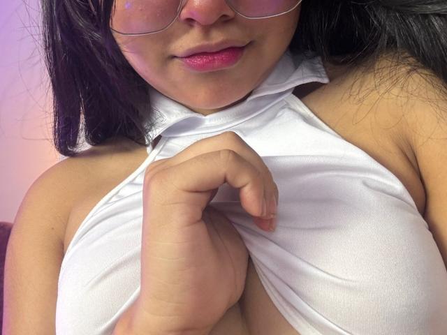 Mei69, avec des lunettes, du rouge à lèvres rose et un haut blanc à enfiler, le tire vers le bas pour exposer ses seins volumineux et ronds. Sa main agrippe le haut, révélant sa poitrine..