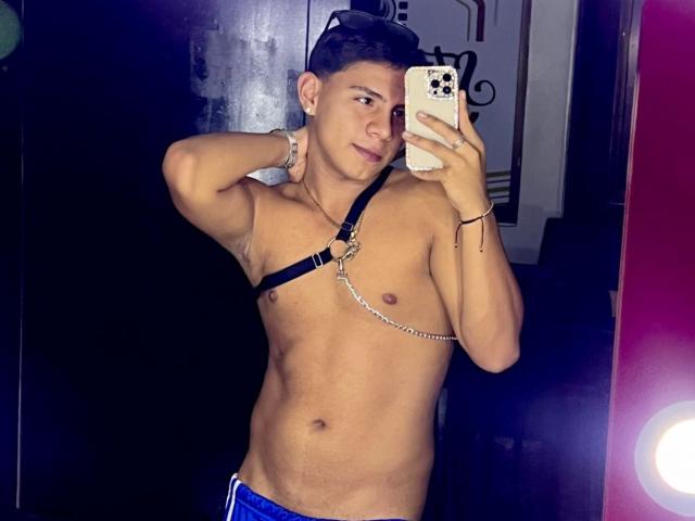 RichiiHott is a muscular, tan-skinned guy with short dark hair, wearing a black leather chest harness, holding a phone, and looking in a mirror, showing his abs.