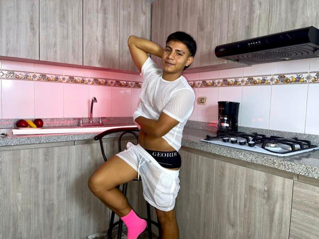 RichiiHott stands in a kitchen, wearing a white, semi-transparent t-shirt and white shorts with "CPD" on the waistband. He has dark skin, short black hair, and a slim, toned build. He's leaning on a black barstool with one hand behind his head, and the other resting on his hip. He's wearing pink socks and has a confident expression. The kitchen has gray wood cabinets, a granite countertop, and a gas stove with a black range hood.