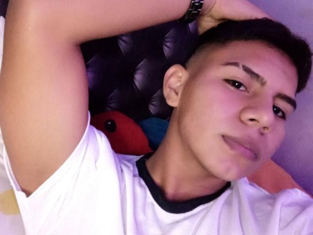 RichiiHott with short black hair, raised arm, white t-shirt, and black watch, lying down, smirking, nose ring, and slightly arched eyebrows, looking at camera.
