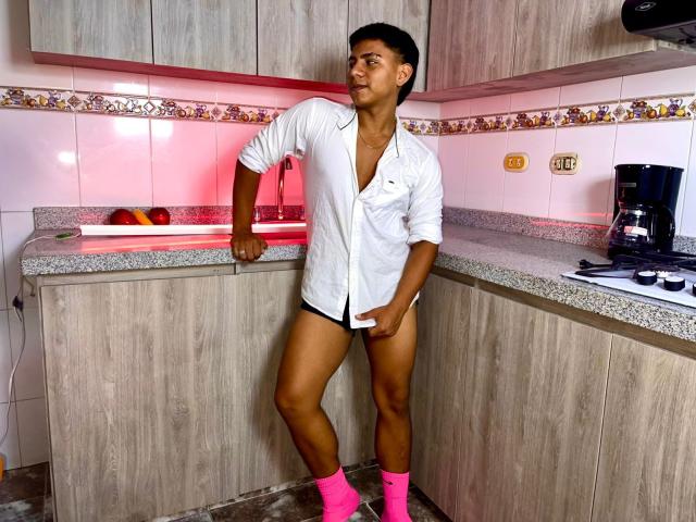RichiiHott stands in a kitchen, wearing an open white shirt, pink socks, and black underwear, with a hand on his exposed cock. He's muscular, tan-skinned, and has short black hair.