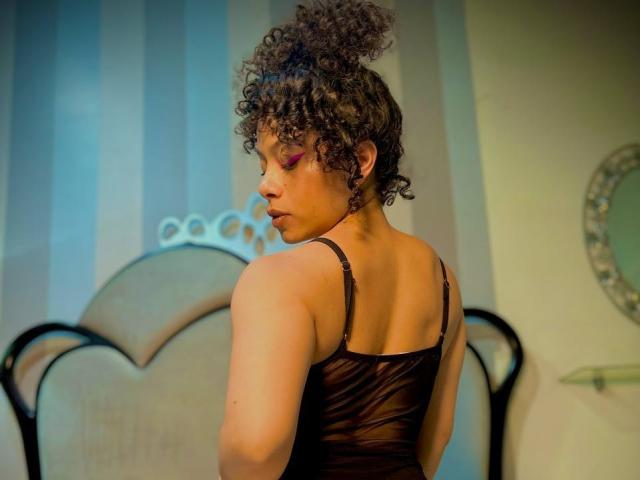 IvannaAndFriend stands with her back to the viewer, head turned slightly to the left. She has curly brown hair in a high bun, wearing a sheer black camisole. Her makeup is vibrant with pink eyeshadow. The background features a striped wall and a decorative headboard. The lighting is warm, highlighting her brown skin and muscular build. Her expression is neutral. The image has a soft, intimate feel.