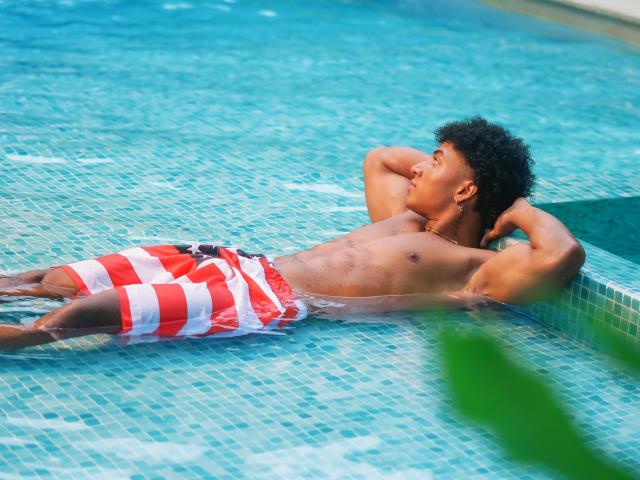 MagnusKing, muscular and tan, with curly black hair, is lounging in a blue-tiled pool, wearing red and white striped swim trunks, arms behind his head, looking to the side.