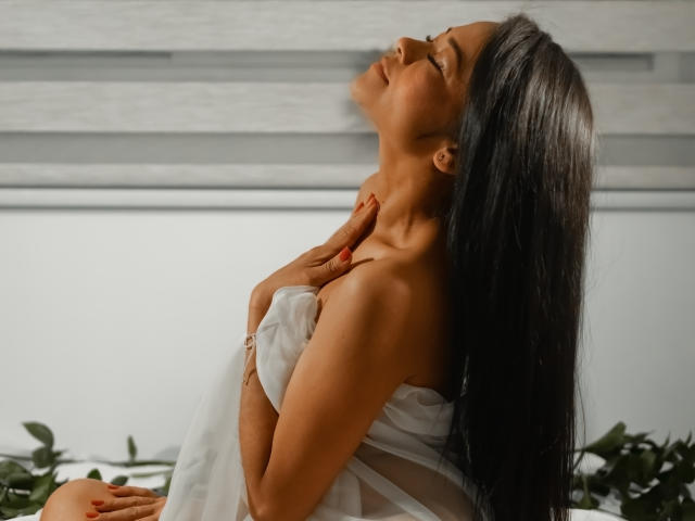 AnelJones, avec de longs cheveux noirs, les yeux fermés, la tête penchée en arrière, les mains sur la poitrine, portant une robe blanche transparente, assise, exposant de petites seins, ongles rouges, plante verte en dessous..