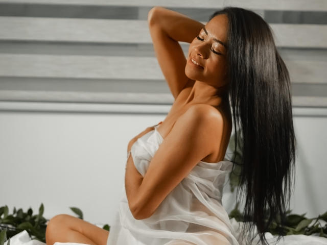 AnelJones, avec de longs cheveux noirs, les yeux fermés, souriante, les bras levés, cachant ses seins avec une robe blanche transparente, assise sur un lit avec des feuilles vertes..