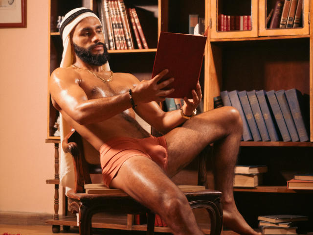Tahiel Foster, un homme musclé, à la peau foncée et avec une barbe, porte un foulard blanc et noir, un short orange, et lit un livre rouge tout en étant assis sur une chaise en bois..