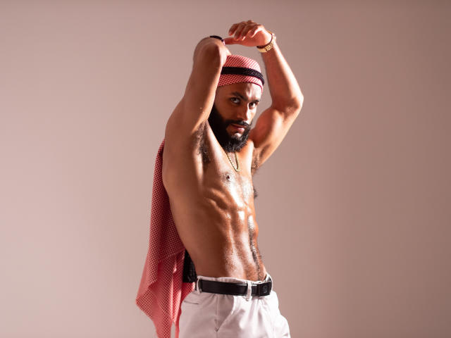 TahielFoster, sin camisa, musculoso, piel oscura, pañuelo rojo en la cabeza, pantalones blancos, brazos levantados, piel brillante, con barba, expresión seria, pulsera dorada, abdominales visibles, de pie, iluminación de estudio..