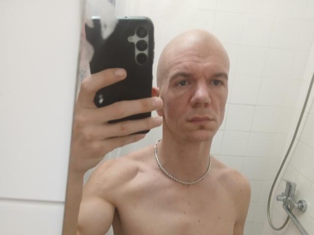 Shirtless HumanTease69 with a bald head, holding a black phone, wearing a silver chain, in a bathroom mirror selfie. His face is slightly puffy, and he's looking serious.