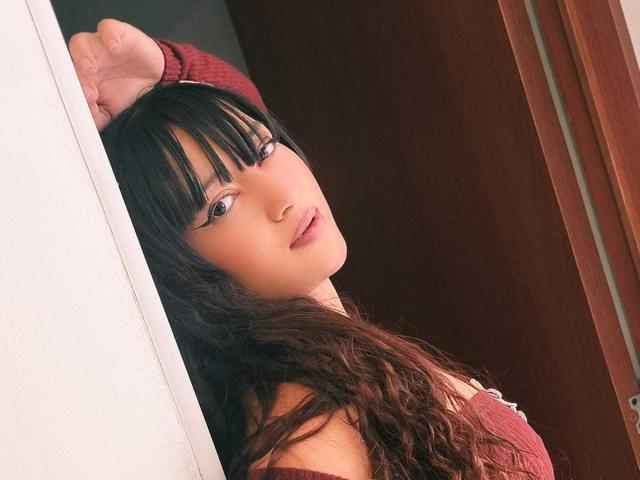 NicoleMartins with black bangs, long wavy hair, and red off-shoulder top, leaning against a white wall, looking back with a slight smile, right arm raised.