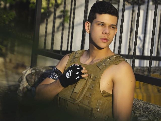 MarcoSullivan with short dark hair, brown eyes, wearing a tan tactical vest, black fingerless glove with white skull, standing by a black metal fence, holding vest straps.