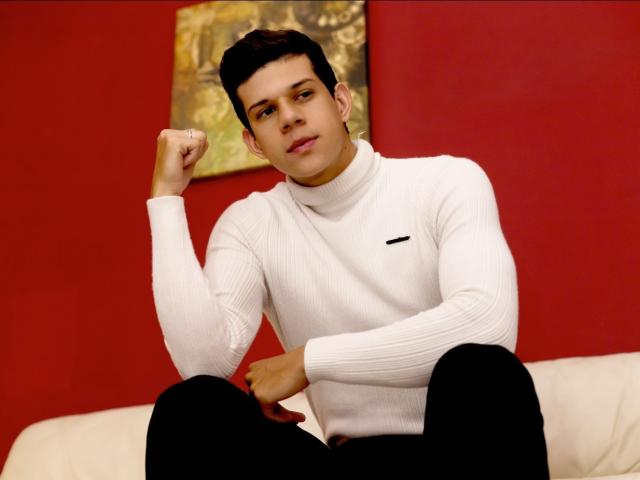 MarcoSullivan, with short black hair, wearing a white turtleneck and black pants, sits on a white couch, flexing his right arm, looking relaxed, with a slight smile.