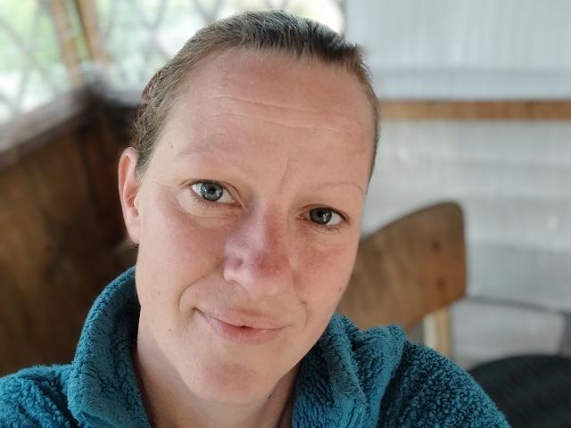 LussyKlara with light skin, blue eyes, and brown hair in a blue fuzzy robe, smiling slightly at the camera, showing small wrinkles on her forehead.
