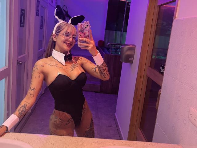CassieInk in a black bunny suit, tattoos covering her arms and chest, white wrist cuffs, black bunny ears, holding phone in mirror, purple bathroom lights, smiling, red lipstick.