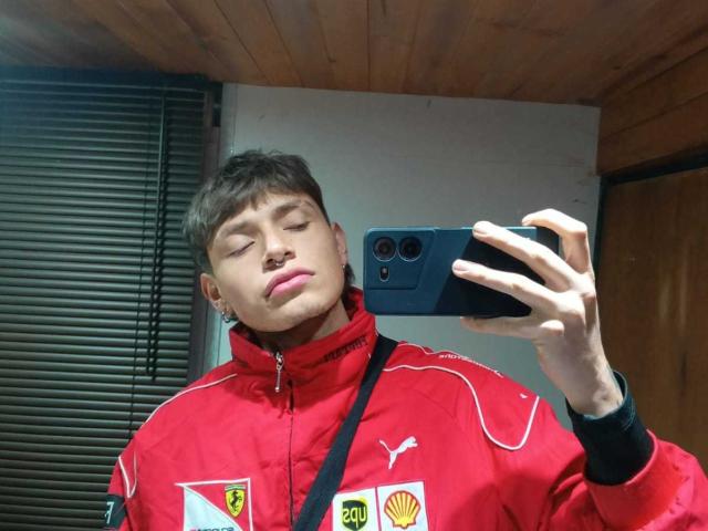 MiaAndDemian with short brown hair, nose ring, and closed eyes, wearing a red Puma jacket, taking a mirror selfie with a blue phone. Her lips are slightly pouted.