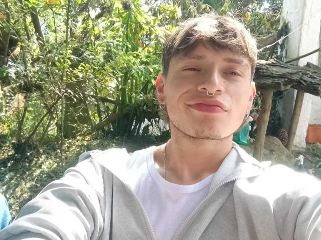 MiaAndDemian with a nose ring, short brown hair, light skin, wearing a gray hoodie over a white tee, smirking, outdoors with greenery in the background.