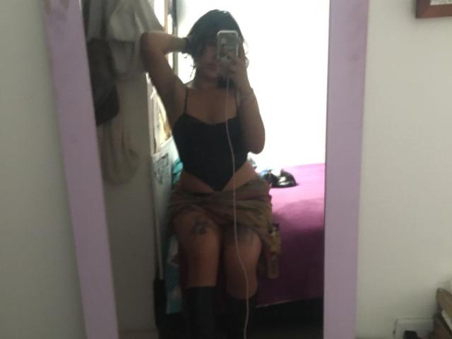 MiaAndDemian with dark curly hair, wearing black lace-up bra and green skirt, sitting on bed, holding phone, showing thigh tattoo, in mirror selfie, with black boots.