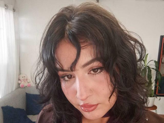 MiaAndDemian has curly black hair, brown eyes, and a nose piercing. She's looking at the camera with a slightly pouty expression, wearing a brown top. Her lips are pierced.