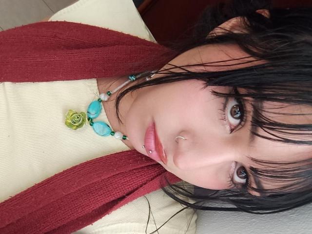 MiaAndDemian with dark wet hair, blue eyes, nose piercing, and pink lip ring, wearing a red cardigan, white top, and turquoise necklace, lying down.