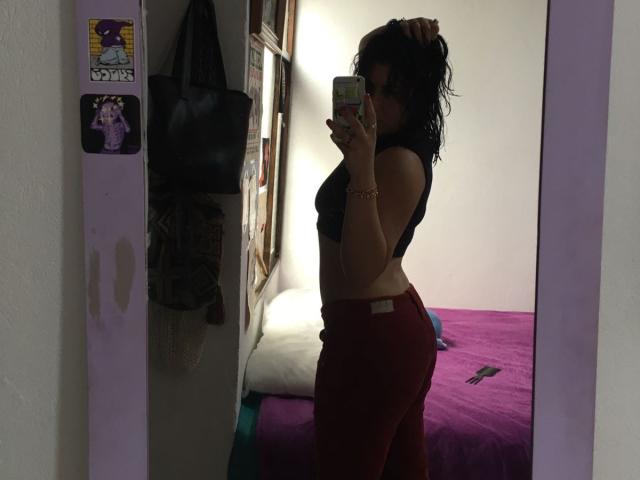 MiaAndDemian with wet black hair, wearing a black top and red pants, holding a phone in a mirror selfie, showing her ass and small tits, in a dimly lit room.