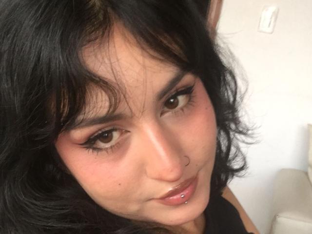 MiaAndDemian has dark wavy hair, brown eyes, black eyeliner, a nose ring, and a lip piercing. She's smiling with a slight pout, wearing a black top.