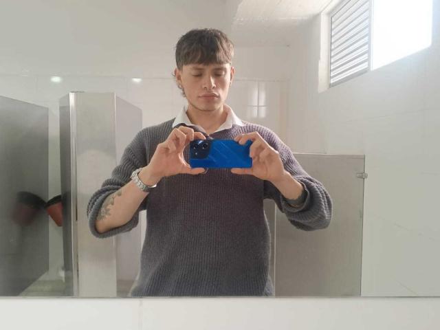 MiaAndDemian with short brown hair, wearing a gray sweater, holding a blue camera, looking down, tattoo on left forearm, standing in a white room, reflection in mirror.
