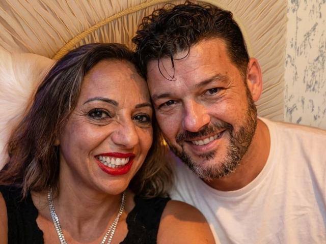 CoupleHottX69 with red lipstick, black dress, and pearl necklace, smiling at camera, next to bearded man in white shirt, both looking happy, wet hair on man.