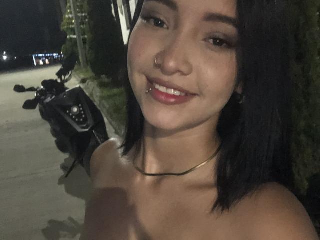 CaritoHam with black hair, nose ring, lip piercing, smiling, bare shoulders, subtle necklace, standing next to a black scooter at night.