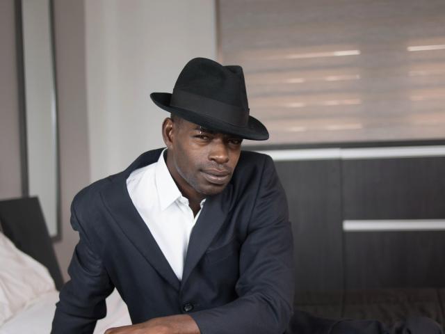TaironTuck is a dark-skinned man with a serious expression, wearing a black fedora and a dark navy suit with a white shirt. He sits on a bed with white pillows, leaning forward. The background has a blurred, horizontal pattern, possibly blinds. The image has a sharp focus on TaironTuck, with a soft, out-of-focus background. The lighting is even, highlighting his face and suit. He looks directly at the camera.