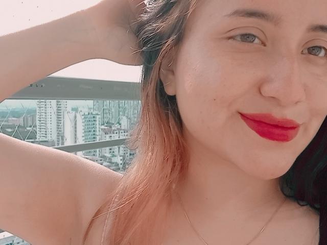 GabrielaMora com batom vermelho, sorrindo, mão no cabelo, pele clara, cabelo castanho, brincos pequenos, colar, seios visíveis, fundo com paisagem urbana, selfie, casual, confiante..