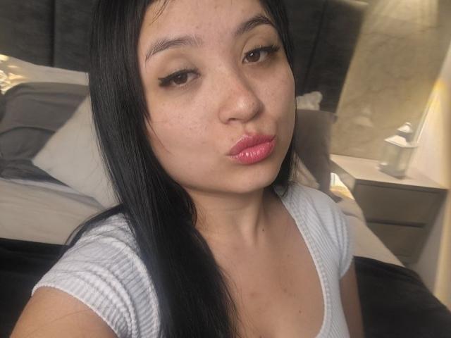 LunaBellaHotty with black hair, light brown skin, and pink lipstick, pouting her glossy lips, wearing a gray ribbed top, in a bedroom setting. Her small tits are visible.