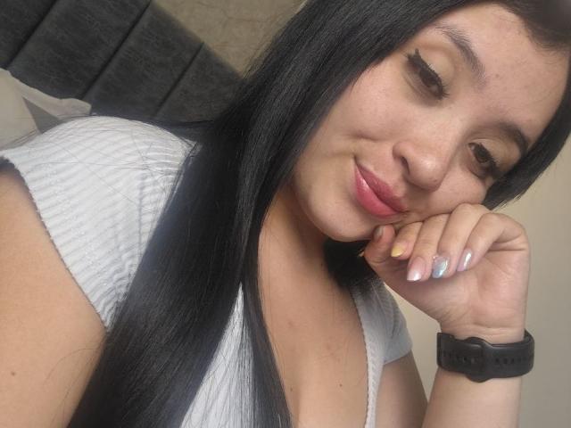 LunaBellaHotty with long black hair, pink lips, and subtle smile, wearing a grey ribbed shirt, resting her chin on her hand with silver nails, and a black watch.