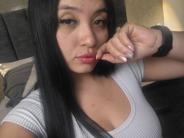 Close-up photo of LunaBellaHotty. She has long, straight black hair, slightly parted lips with glossy pink lipstick, and light brown eyes with subtle eyeliner. She's wearing a light gray, ribbed, short-sleeve top with a low neckline, showing a hint of cleavage. Her right hand is near her mouth, fingers slightly curled, with a black smartwatch on her wrist and nails painted in a light, glittery color. Her skin is light with a few small freckles on her cheeks. The image has a natural, slightly overexposed look.